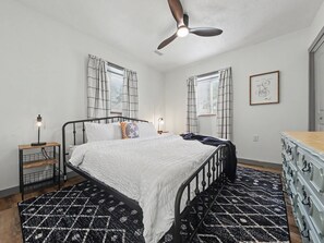 A bedroom with a bed, a ceiling fan, a nightstand with a lamp, a dresser, and a patterned rug.