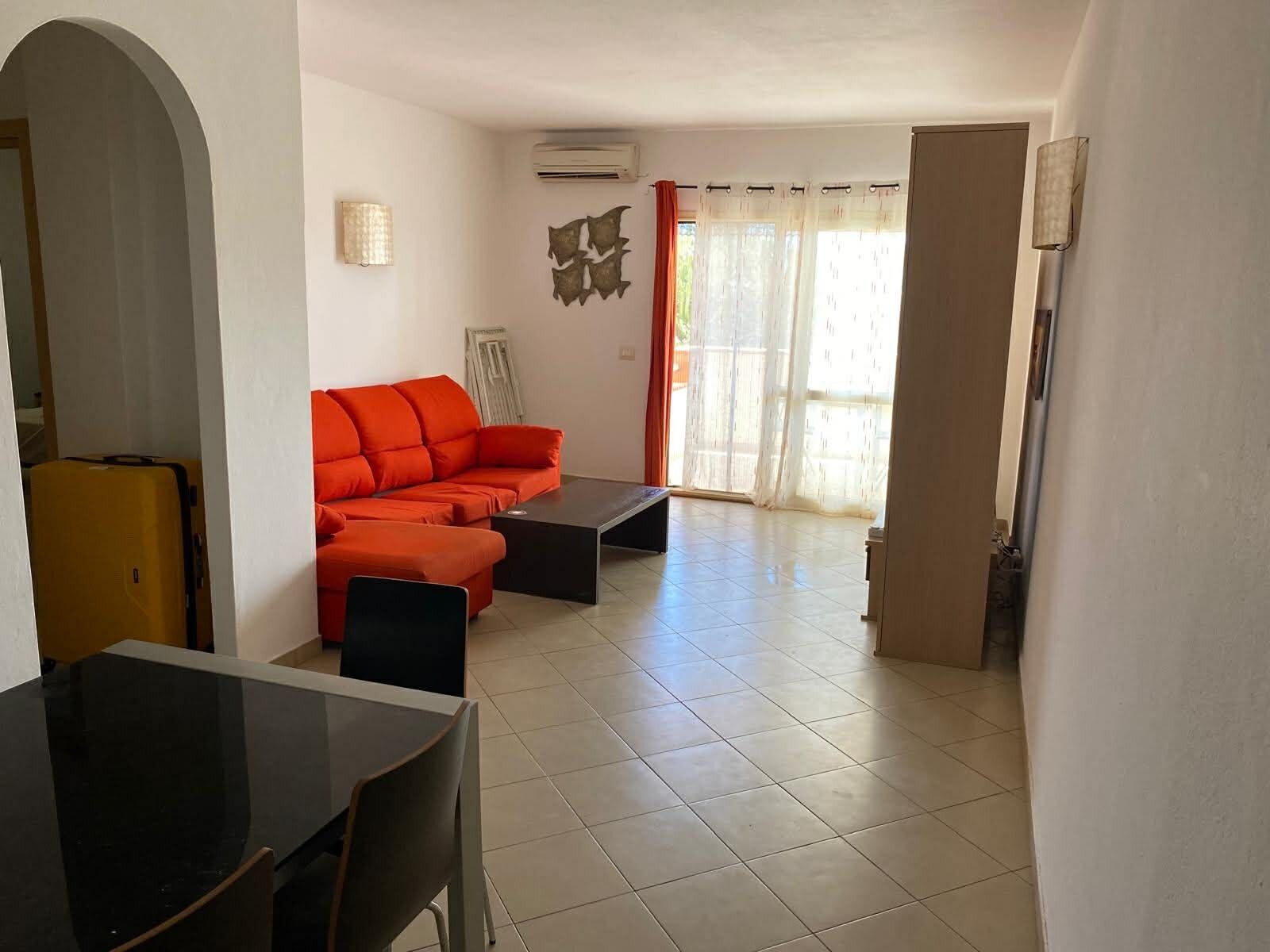 A living room with a sofa, a dining table, and chairs. There is a sliding glass door leading to a balcony.