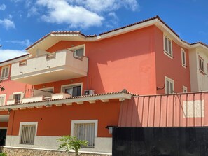 A multi-story residential building with balconies, a stone wall in front, and a clear blue sky. - Peaceful 2-bedroom apartment with AC in Santa Maria. 400m from the beach. (Santa Maria)