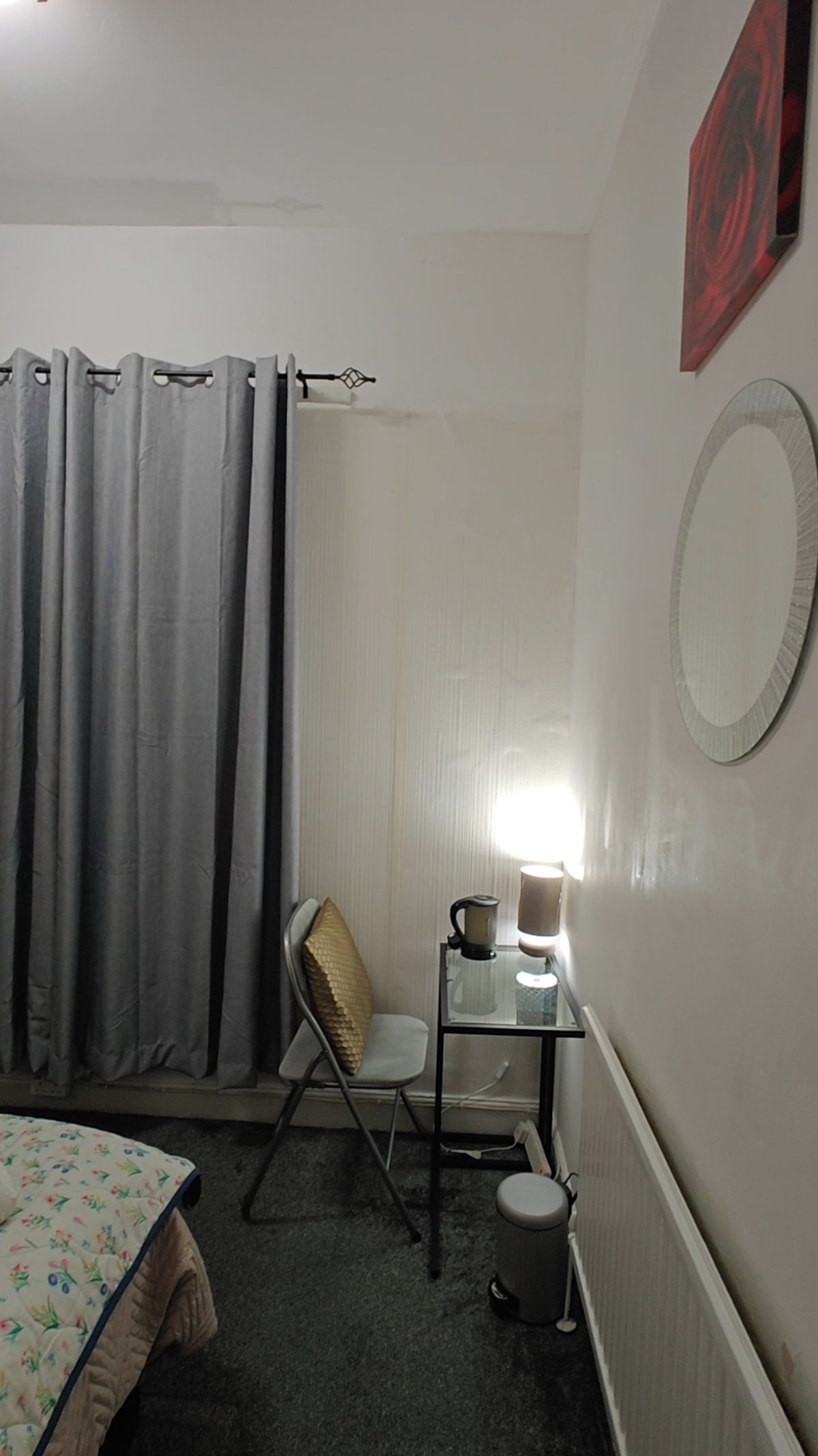 A small, compact room with a bed, a chair, a table with a lamp, a radiator, and a curtain.