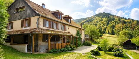 A two-story house with a covered porch, a stone chimney, and a hillside view.