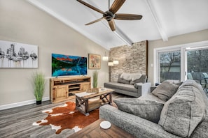 A living room with a ceiling fan, a large painting, a stone wall, a gray sofa, and a wooden coffee table. - Hestia: Remodeled 3 Bed gem near downtown (Austin)
