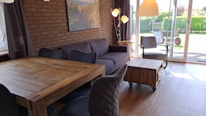 Ein modernes Wohnzimmer mit Holztisch, Sofa und Stühlen, einem großen Fenster mit Blick ins Freie und einer Stehlampe.
