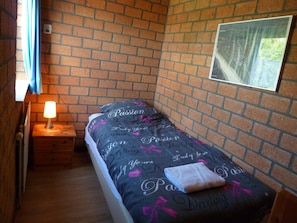 Ein kleines Zimmer mit einem Einzelbett, einem Nachttisch mit Lampe, einer Backsteinwand und einem gerahmten Bild.