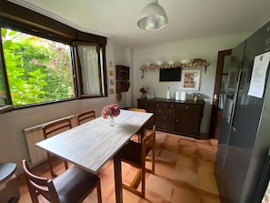Ein Essbereich mit Tisch, Stühlen, einem Blumenstrauß, einem Fenster mit Vorhängen sowie eine Küchenzeile mit Schränken und einem Kühlschrank.