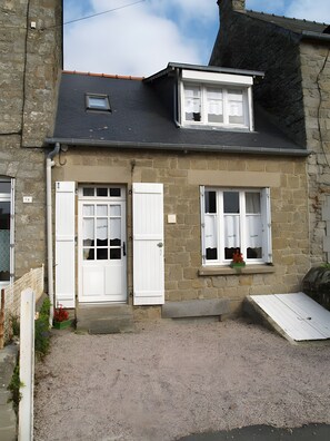 A two-story stone building with a gravel driveway, white doors, and a small window with a flower box. - House 'La Rue, Petite Maison de Pêcheur proche plage' (Cancale)