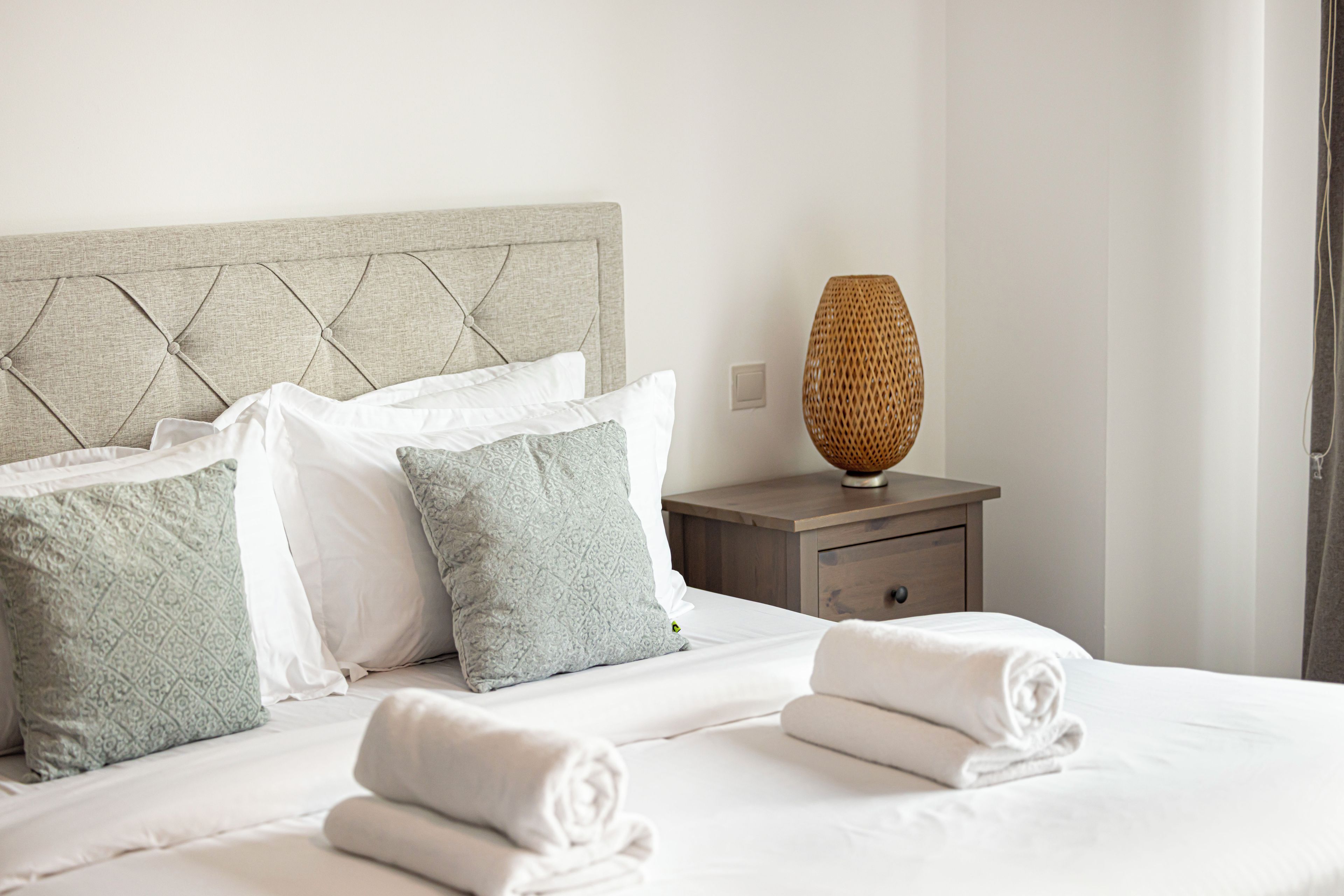 Una cama bien hecha con sábanas blancas y toallas enrolladas, una mesita de noche de madera con una lámpara de mimbre y un cabecero con diseño acolchado.