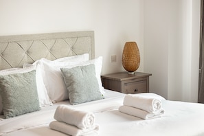 Een keurig opgemaakt bed met witte lakens en opgerolde handdoeken, een houten nachtkastje met een geweven lamp, en een hoofdbord met een gestuufd design.