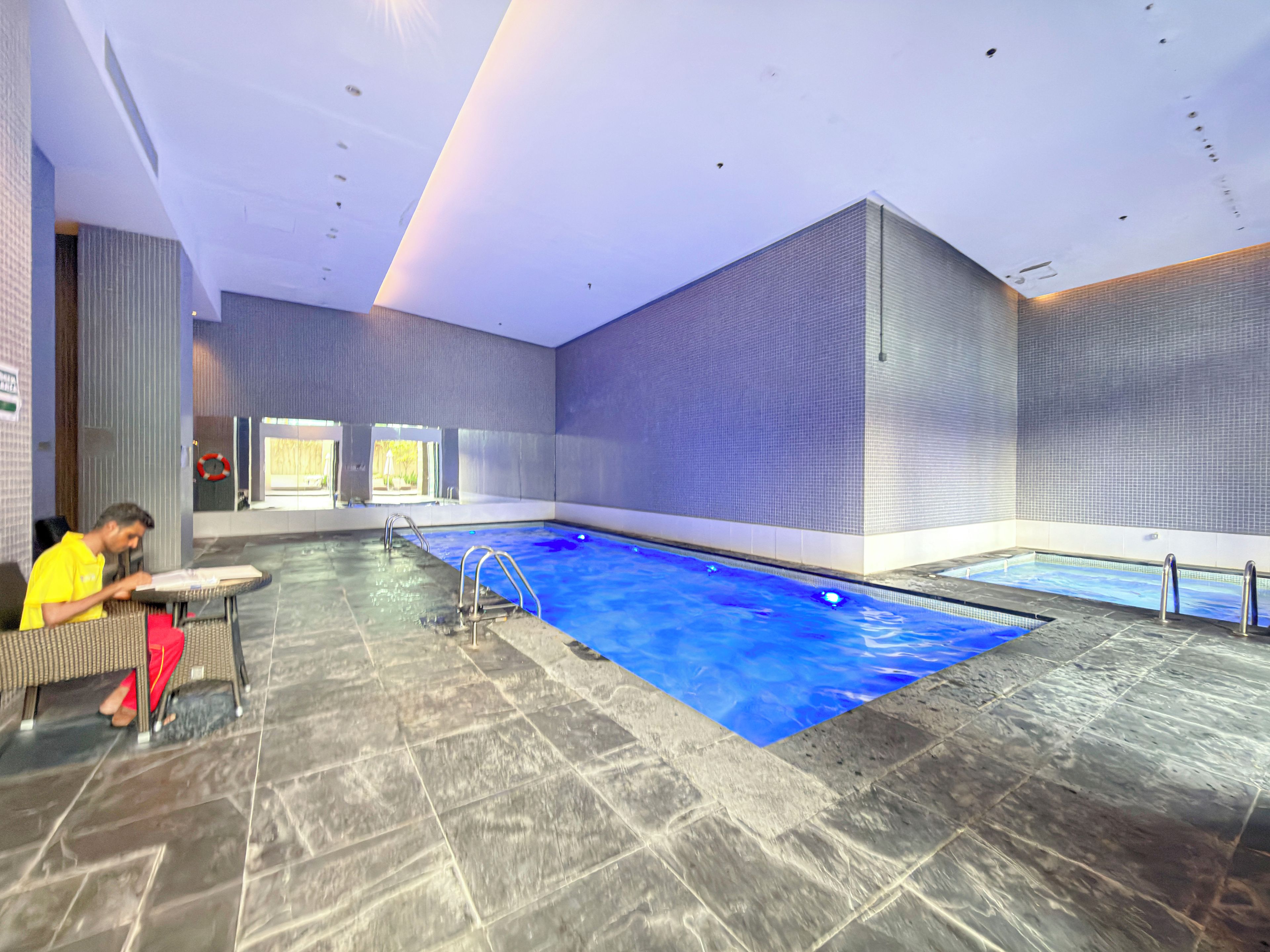 Piscina interior com piso ladrilhado, uma escada e uma pessoa sentada à mesa.