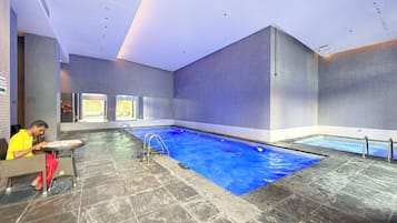 Piscina interior com piso ladrilhado, uma escada e uma pessoa sentada à mesa.