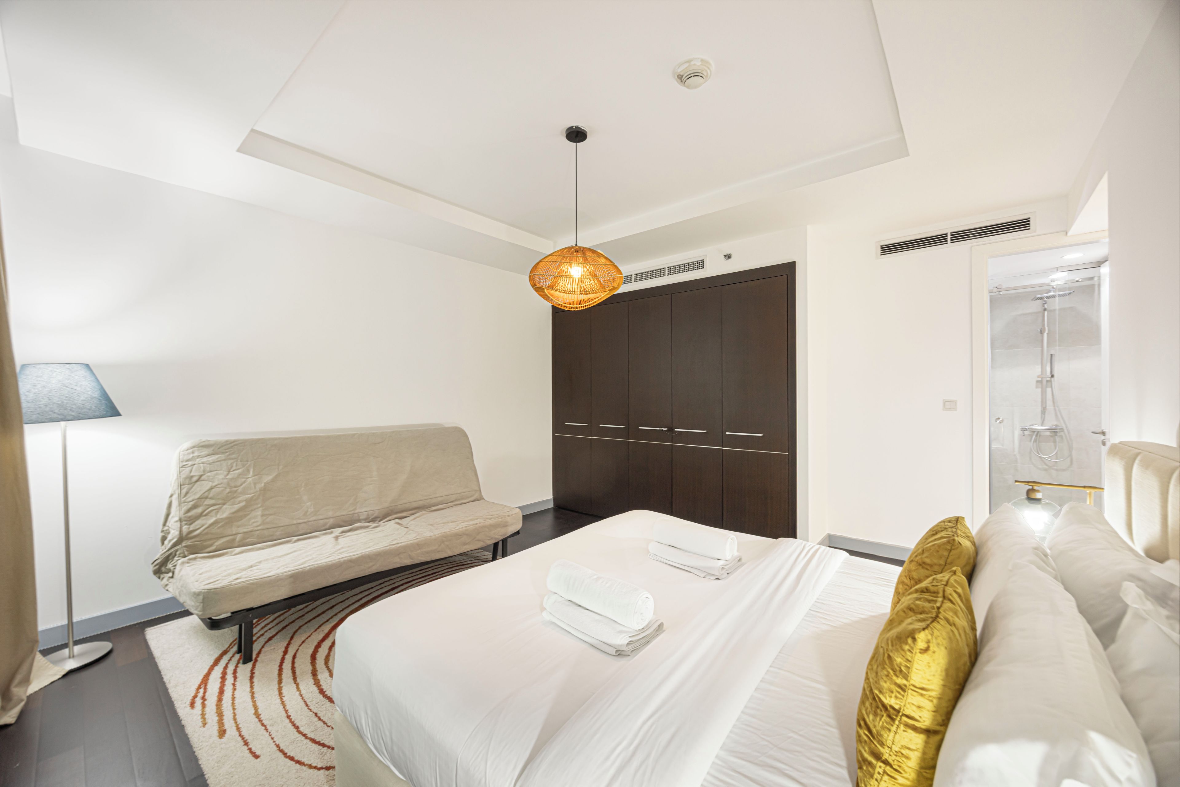 A hotel room with a bed, a sofa, a hanging light fixture, a wardrobe, and a shower area.