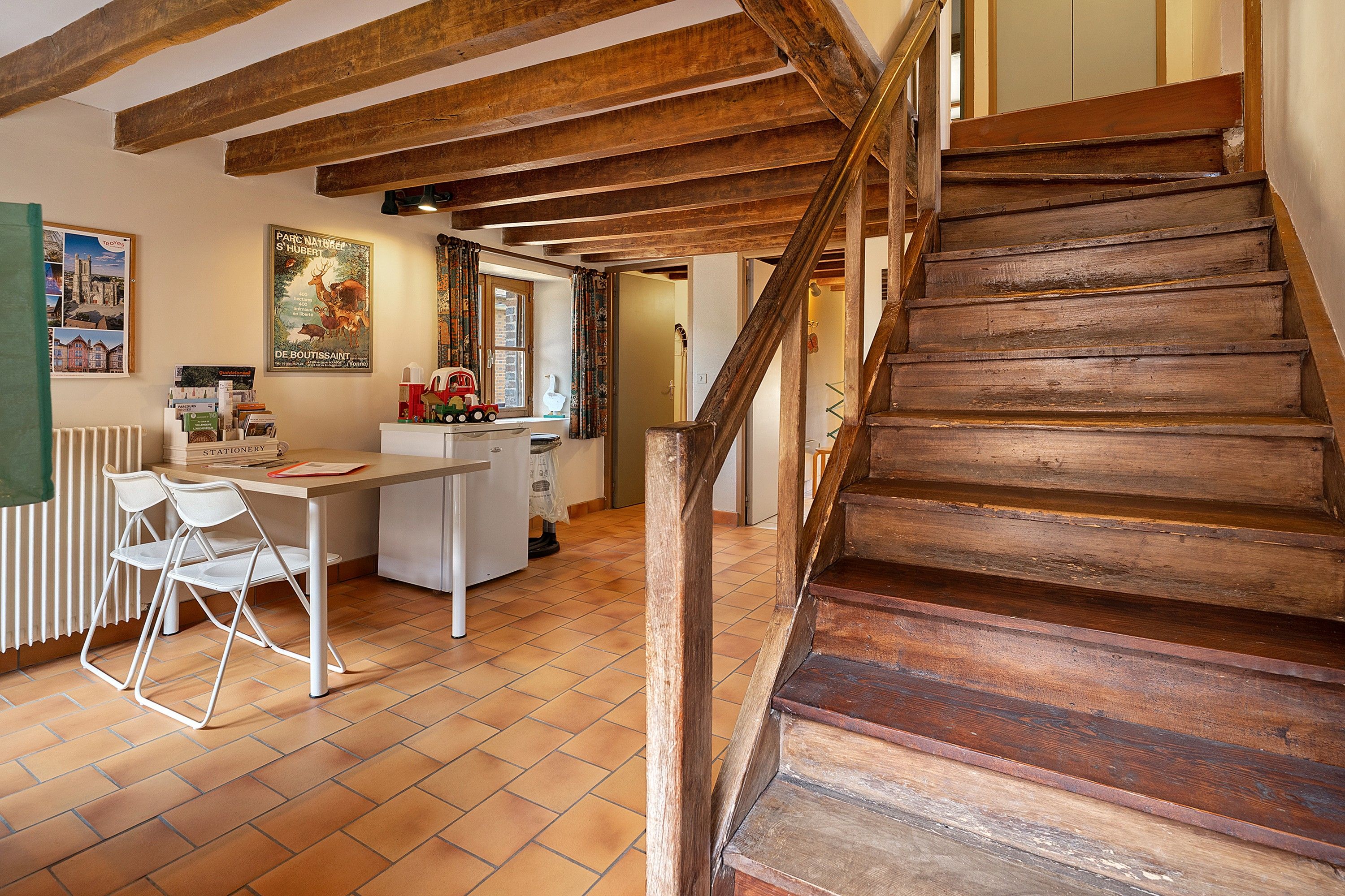 Un interior rústico con una escalera de madera, un comedor con mesa y sillas, y una cocina visible a través de un umbral abierto.