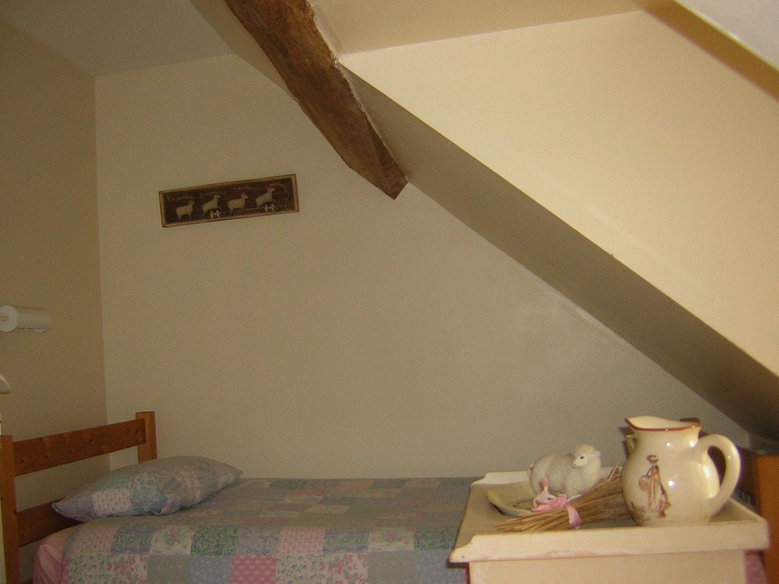 Ein Schlafzimmer mit hölzernem Kopfteil, einer gesteppten Bettdecke, einem kleinen Nachttisch mit einem Keramik-Gießkännchen und einem dekorativen Gegenstand sowie einem gerahmten Bild an der Wand.