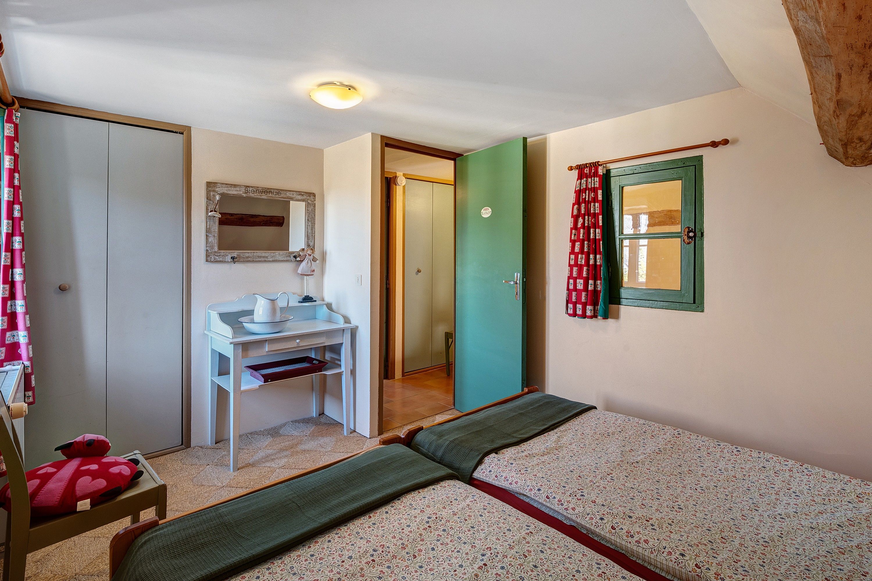 Une pièce comprenant un lit, un petit bureau avec un miroir, une porte et une fenêtre avec des rideaux.