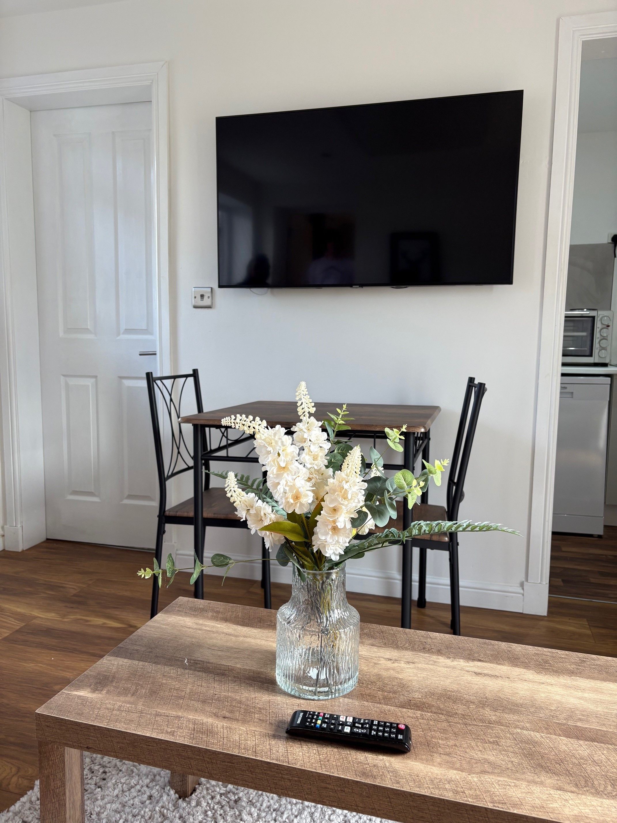 A living room with a wooden coffee table, a vase with flowers, a remote control, a television mounted on the wall, and a dining area with chairs.