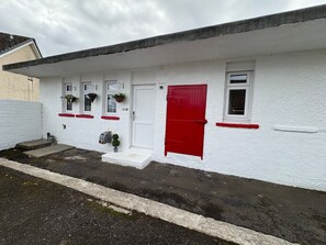 A white building with a red door, windows with hanging flower baskets, and a small step leading to the entrance.