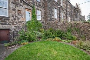 A stone building with a red door, a small garden with grass and various plants, and a stone wall. - Banrigh House - a terrace that sleeps 6 guests in 3 bedrooms (Stirling)