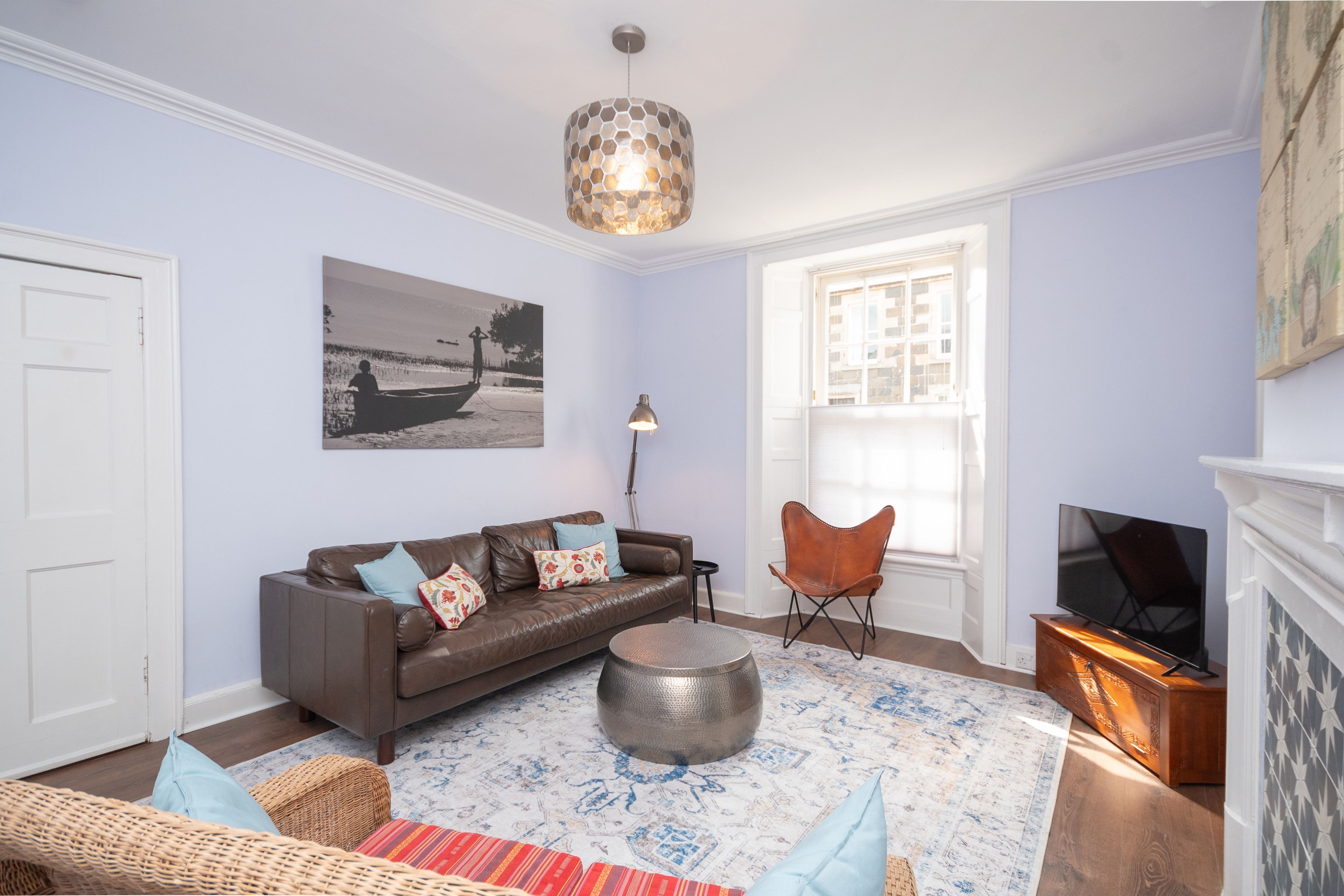 A living room with a brown leather sofa, a round coffee table, a wicker chair, a floor lamp, and a television.