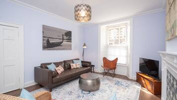 A living room with a brown leather sofa, a round coffee table, a wicker chair, a floor lamp, and a television.