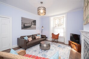 A living room with a brown leather sofa, a round coffee table, a wicker chair, a floor lamp, and a television. - Banrigh House - a terrace that sleeps 6 guests in 3 bedrooms (Stirling)