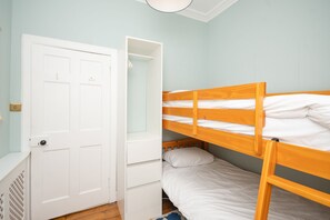 Een stapelbed met een witte matras en een houten frame, een witte deur met een sleutelgat, en een klein wit kastje met lades.