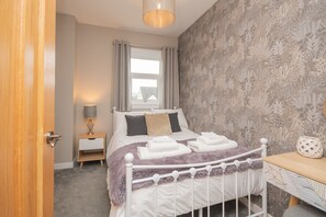 A bedroom with a bed, a nightstand, a lamp, a window with curtains, and a patterned wall. - Cochrane House -  a house  that sleeps 5 guests  in 3 bedrooms (Stirling)