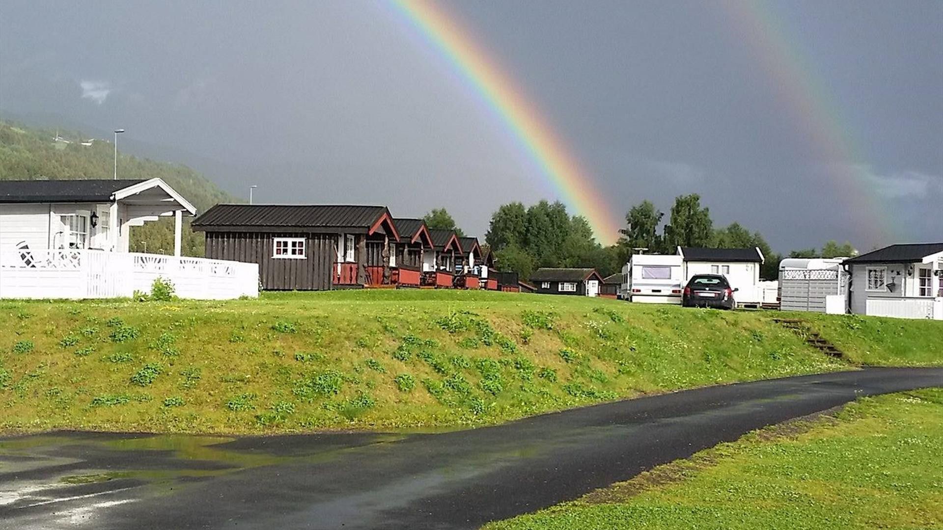 Paesaggio rurale con una strada, un arcobaleno e diversi edifici, tra cui una casa bianca, una struttura con tetto rosso e un camper bianco.