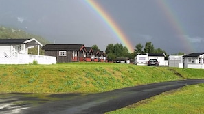 A rural setting with a road, a rainbow, and several buildings including a white house, a red-roofed structure, and a white trailer.