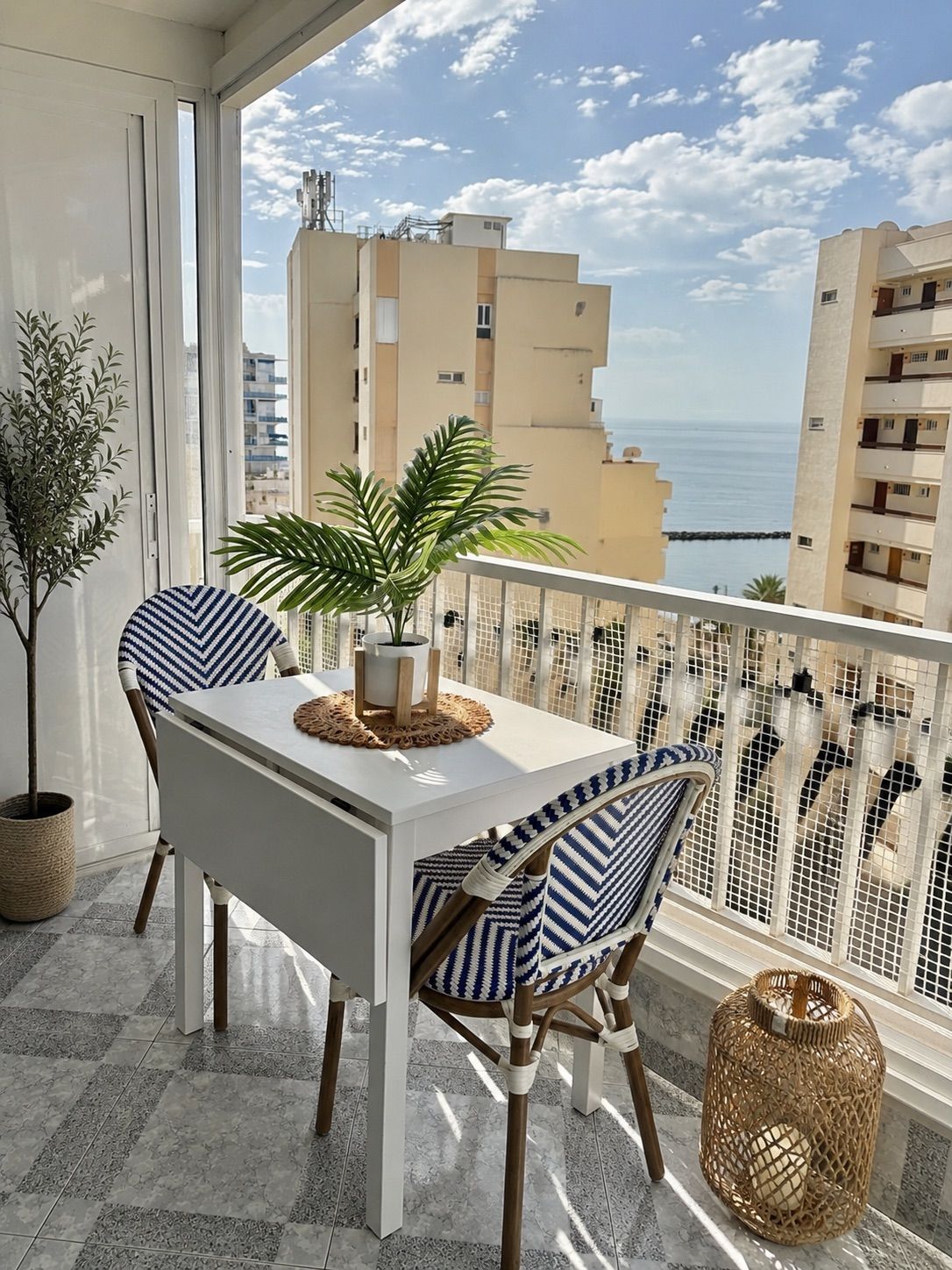 A balcony with a white table and chairs, a potted plant, and a view of the sea and buildings.