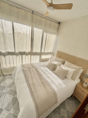 A bedroom with a large bed, a ceiling fan, a nightstand, and a window with sheer curtains. - Apolo Mediterranean Living (Marbella)