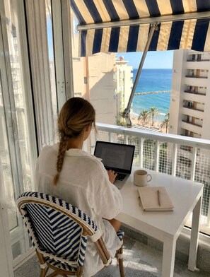 Une personne travaille sur un ordinateur portable, assise à une table blanche, avec vue sur l’océan et les bâtiments.