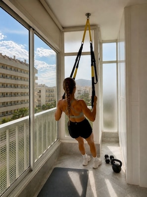 A person using a suspension trainer in a room with large windows, a balcony, and exercise equipment. - Apolo Mediterranean Living (Marbella)