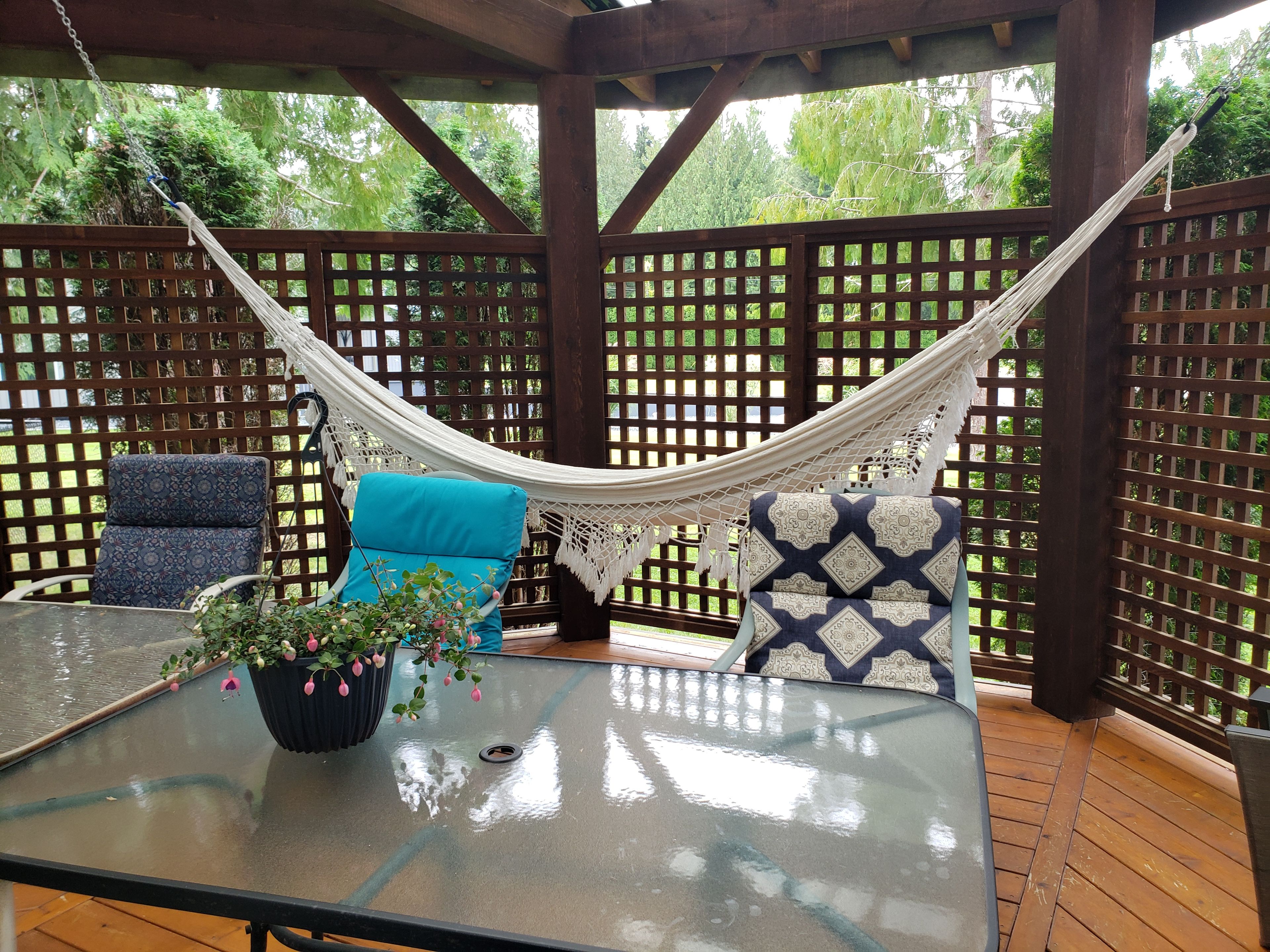 A hammock suspended between wooden lattice panels, a glass table with a potted plant, and two patterned chairs.