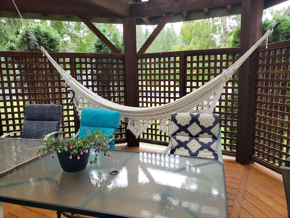 A hammock suspended between wooden lattice panels, a glass table with a potted plant, and two patterned chairs.