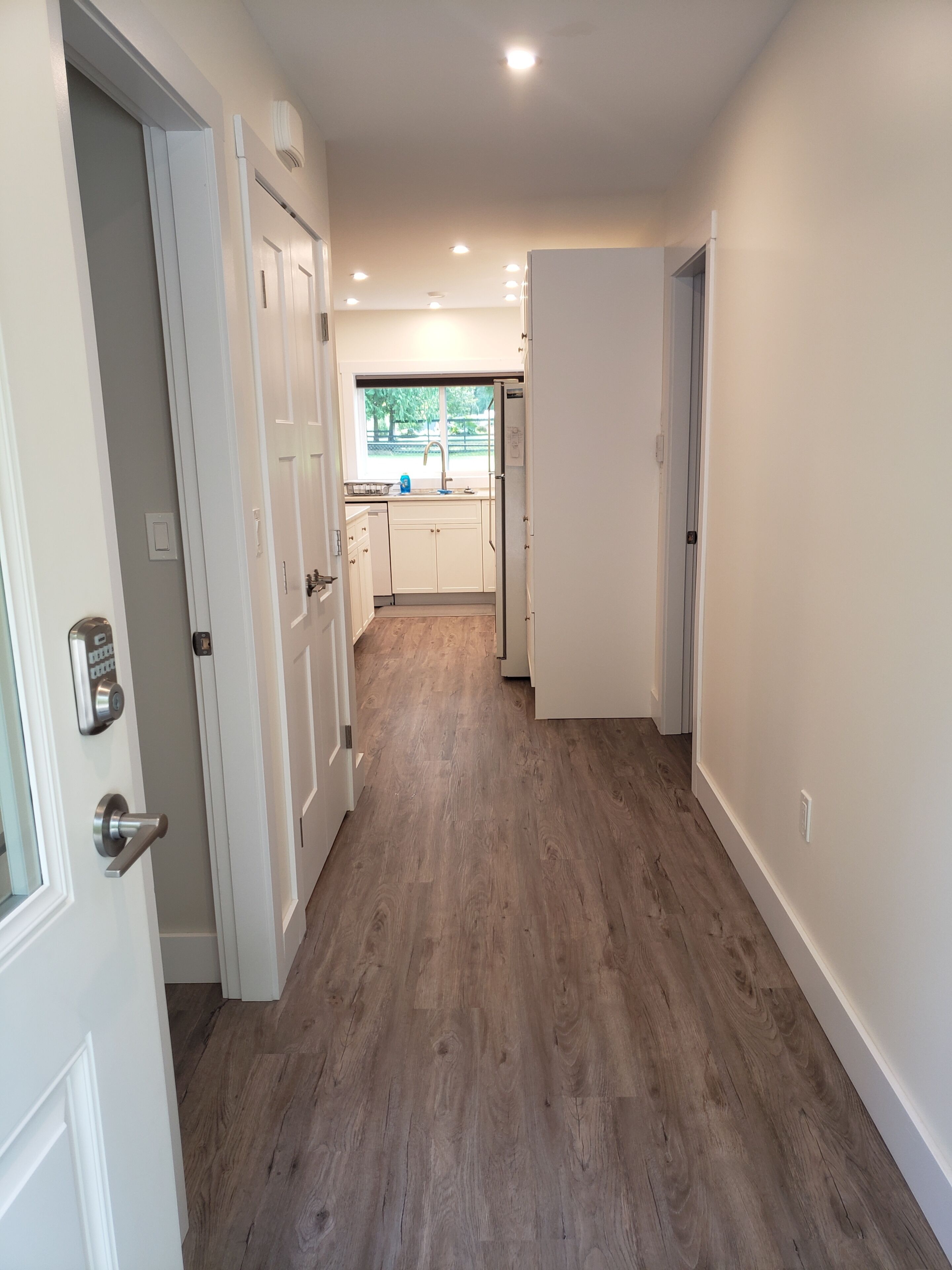Un couloir au plancher de bois menant à une cuisine avec des meubles blancs et une fenêtre.