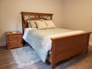 A wooden bed with a headboard, a bedside table with drawers, and a lace throw on the floor. - Quiet, private brand new 1 bdm on Wooded Estate by shops, airport & beach (Powell River)