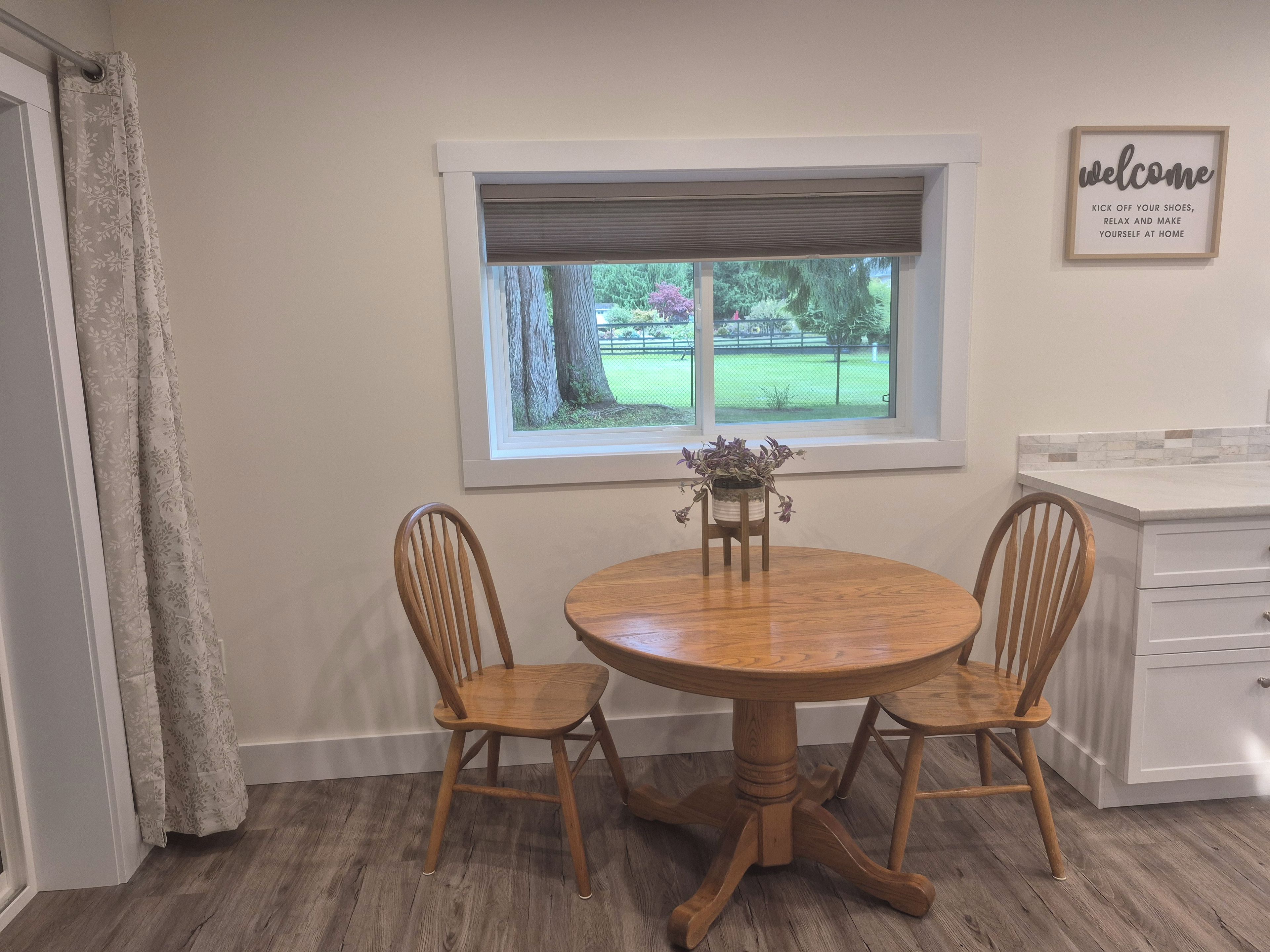Un coin repas avec une table ronde en bois, deux chaises en bois, une fenêtre donnant sur un jardin et un panneau « Bienvenue » accroché au mur.