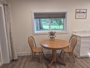 A dining area with a round wooden table, two wooden chairs, a window with a view of a garden, and a 'welcome' sign on the wall.
