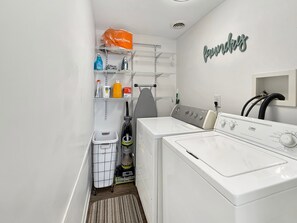 A laundry area with two washing machines, a dryer, a shelf with cleaning supplies, and a wall-mounted rack. - Cozy Vermilion Retreat Near Lake Erie - Sleeps 3 (Vermilion)