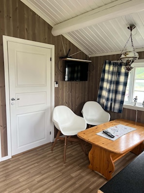 A room with a white door, two white chairs, a wooden table, a television mounted on the wall, and a window with a plaid curtain. - Cosy Camping Cabin no. 12 in Øyer (Øyer)