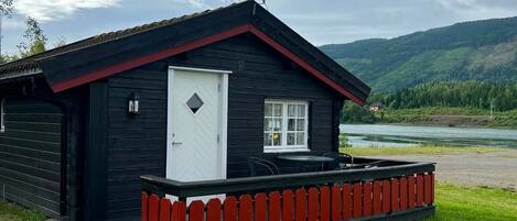 A cabin with a red porch and a white door, surrounded by a grassy area and a river.