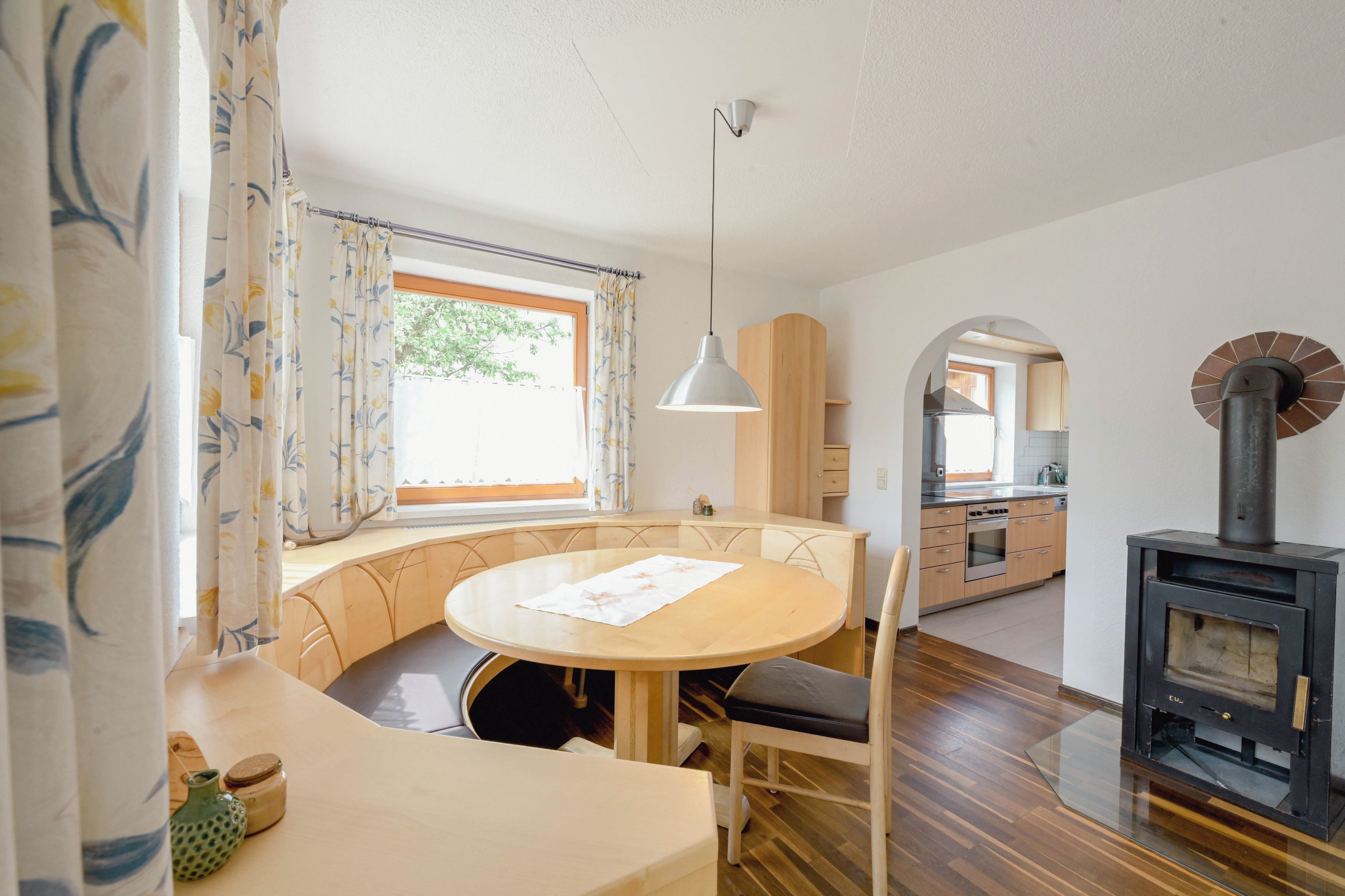 Een gezellige kamer met een ronde houten tafel, een bank, een raam met gordijnen, een keukenhoek en een houtkachel.