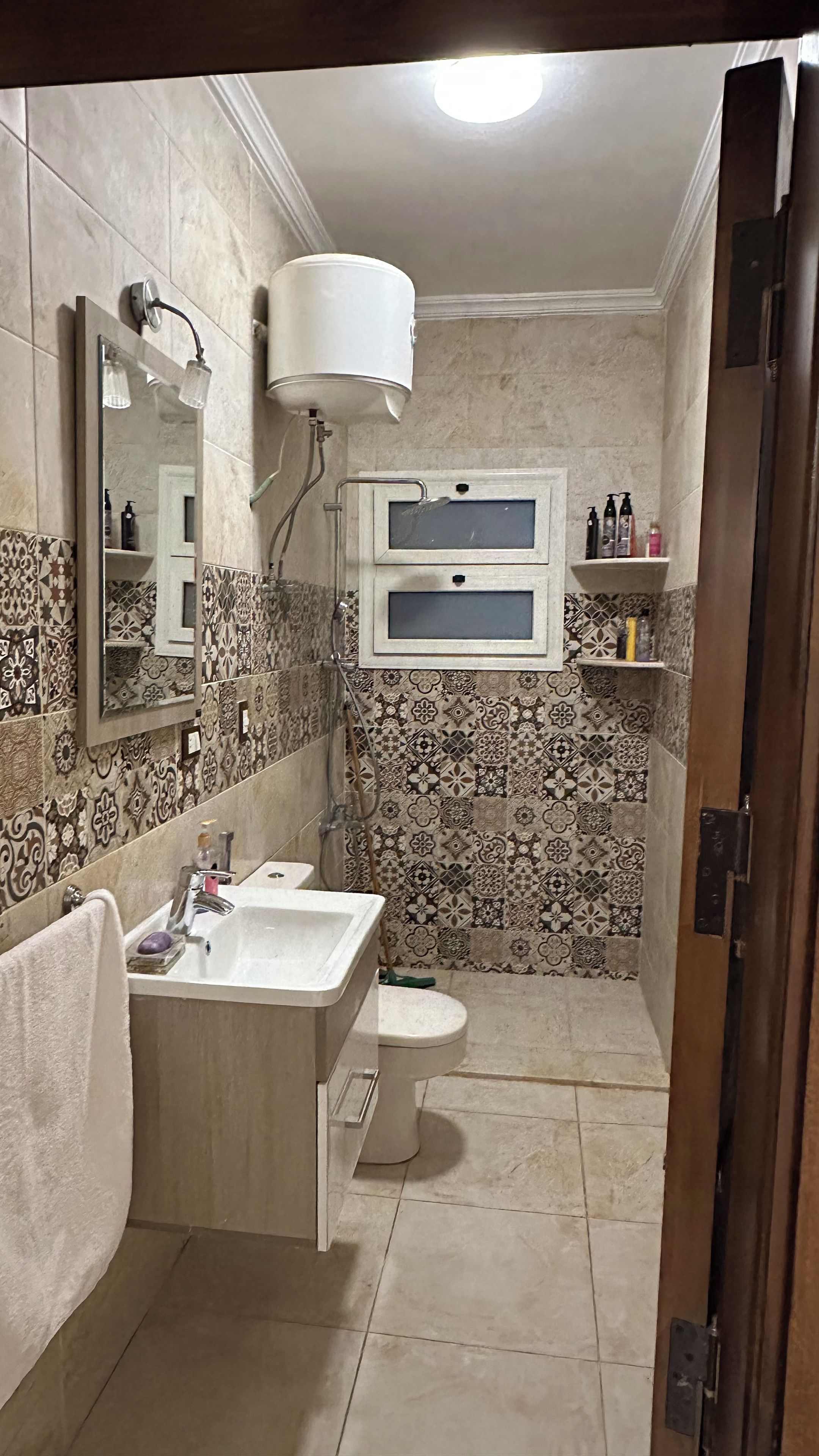 A bathroom with a sink, toilet, and a mirror. There is a showerhead above the sink and a towel hanging on the wall.