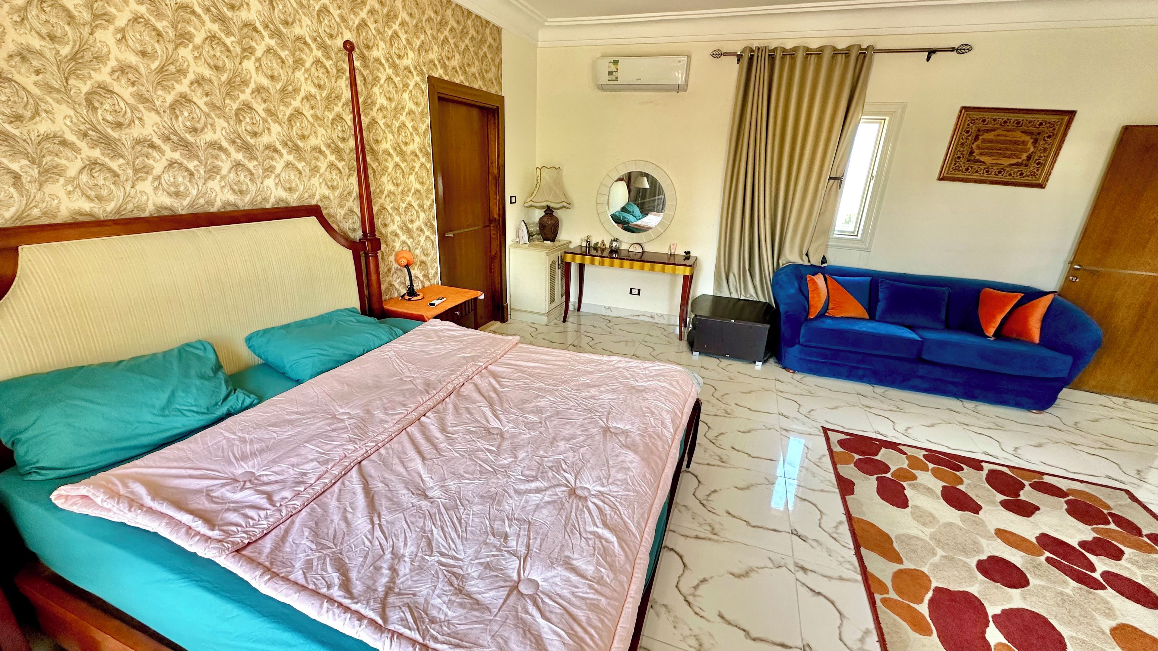 Una habitación de hotel con una cama, un sofá azul, una mesita, un espejo y una alfombra estampada.