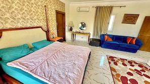 Quarto de hotel com cama, sofá azul, mesa pequena, espelho e tapete estampado.