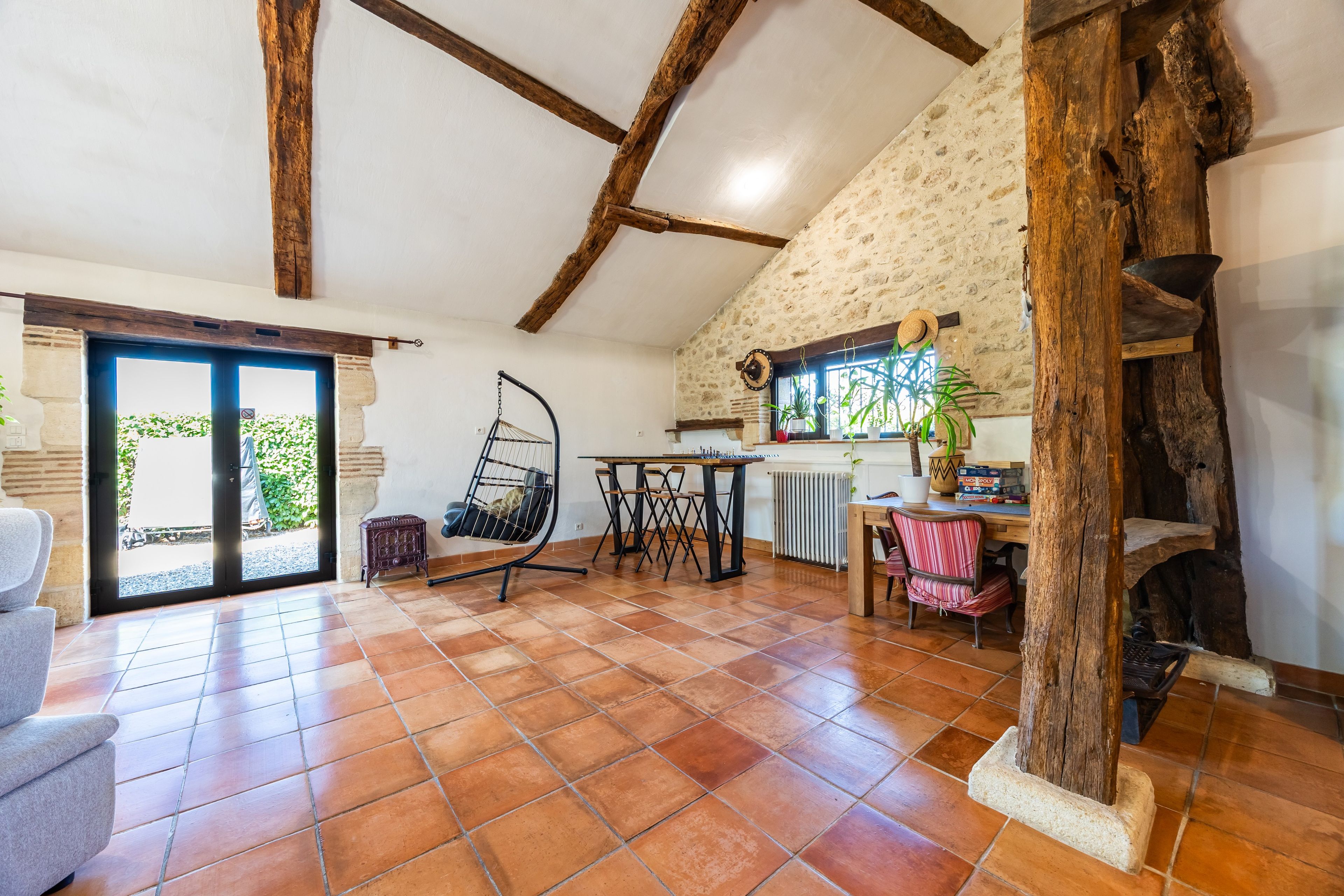 Un'ampia stanza con pavimento piastrellato, soffitto in legno, zona pranzo con sedie, una sedia a dondolo e vista sull'esterno.