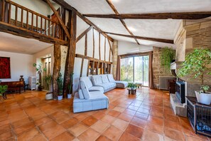 A spacious living room with a high ceiling, exposed wooden beams, a large grey sofa, and a stone fireplace. - Holiday Home 'Domaine Liminalis' with Private Terrace, Garden and Wi-Fi (Saint-Martin-Petit)