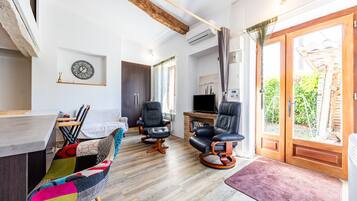 A modern living room with a wooden floor, a television, two black leather chairs, a colorful sofa, and a large window with curtains.