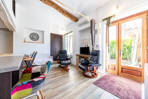 Uma sala de estar moderna com piso de madeira, uma televisão, duas poltronas de couro preto, um sofá colorido e uma janela grande com cortinas.