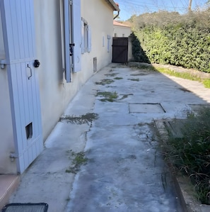 A narrow concrete pathway leading to a building with white doors and shutters.